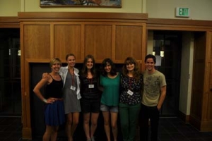 Six people stand together, smiling in front of a wooden wall. They are casually dressed, and some wear name tags. The setting appears to be indoors, possibly a hallway or lobby.