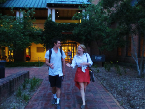 Two people are walking and talking on a brick path during dusk. The person on the left is wearing a white t-shirt and shorts, and the person on the right has a white shirt and red skirt. A building with lights is in the background.