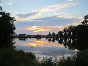 A tranquil lake reflecting the colorful sky at sunset, with silhouettes of surrounding trees and distant buildings. Wispy clouds add texture to the serene scene, framed by greenery in the foreground.