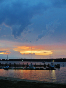 A serene marina at sunset with boats docked at a pier. The sky transitions from deep blue to warm orange near the horizon, reflecting on the calm water. Dark clouds add contrast to the scene.