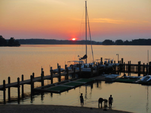 Sunset over a calm lake with a docked sailboat. Three silhouetted people stand in shallow water near the shore. The sky is painted with shades of orange and pink, while the horizon is lined with dark trees.