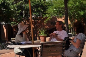 Three people sit at a wooden picnic table under an umbrella, engaged in conversation. One wears a wide-brimmed hat, another a white shirt, and the third a striped shirt. Sunlight filters through trees in the background, casting light shadows.
