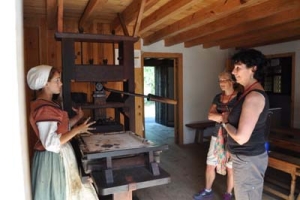 A woman in historical attire demonstrates an old printing press to two visitors in a wooden room. The visitors stand attentively, observing the explanation. The room has wooden beams and subtle natural lighting.