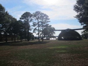 Outdoor stage with a large canopy set in a grassy park. Tall trees surround the area, and a building is visible on the left. A body of water is in the background under a partly cloudy sky.