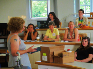 A person with curly hair presents in a classroom, holding a book. Five people sit around a semi-circular desk, listening attentively. A laptop is open in front of one person. Cardboard boxes are on a front table. Natural light comes through windows.