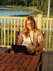 A person with long red hair and glasses sits at a wooden table on a deck, using a Toshiba laptop. They are wearing a tie-dye T-shirt. The background shows a beach, water, and trees under a clear blue sky.