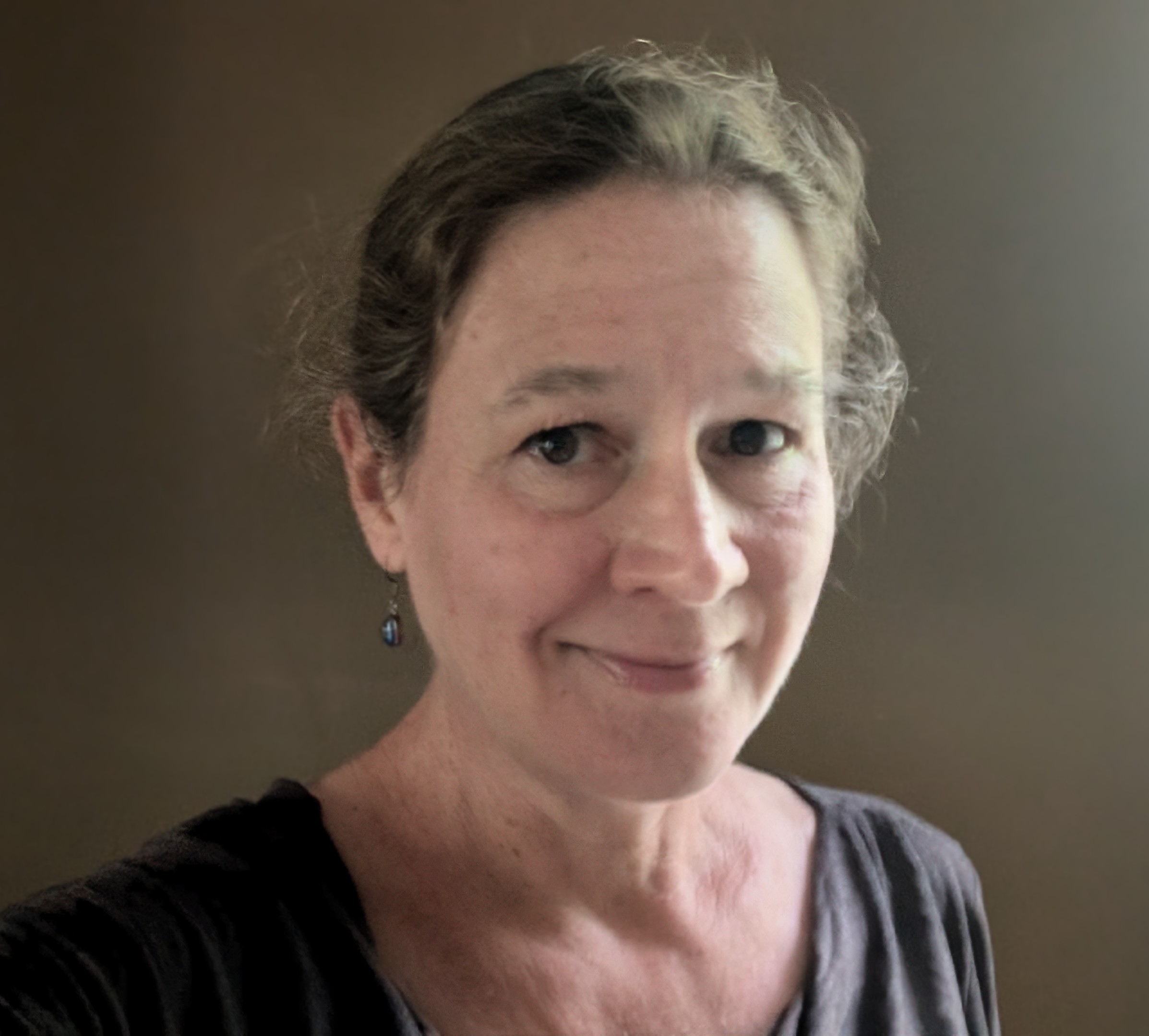 Mary Quade Author Photo 2024 A person with light brown hair pulled back wearing a dark top and an earring, smiles softly at the camera. The background is a plain grayish wall.