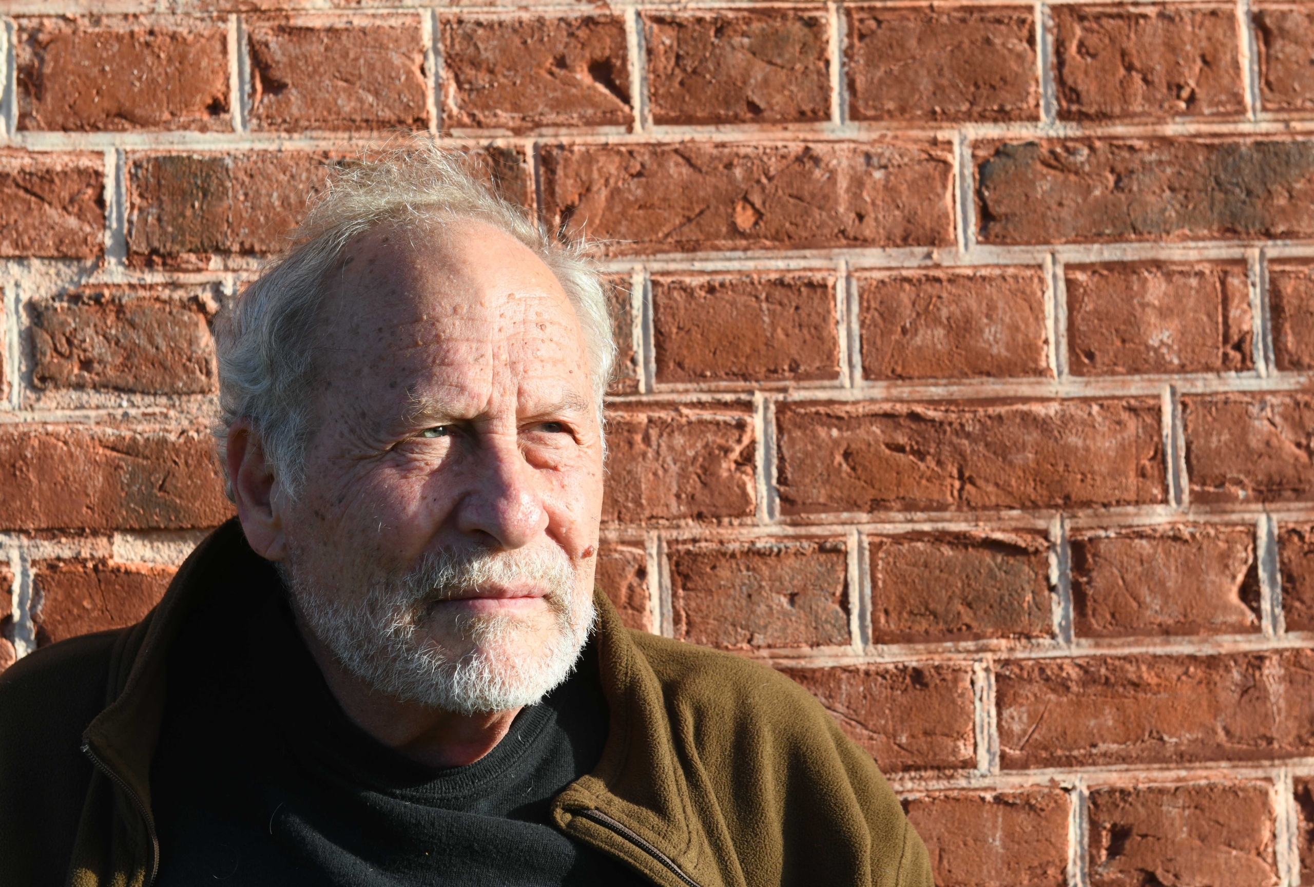 Wayne Karlin Older man with a beard and wearing a brown jacket stands in sunlight against a red brick wall, gazing thoughtfully to his left.