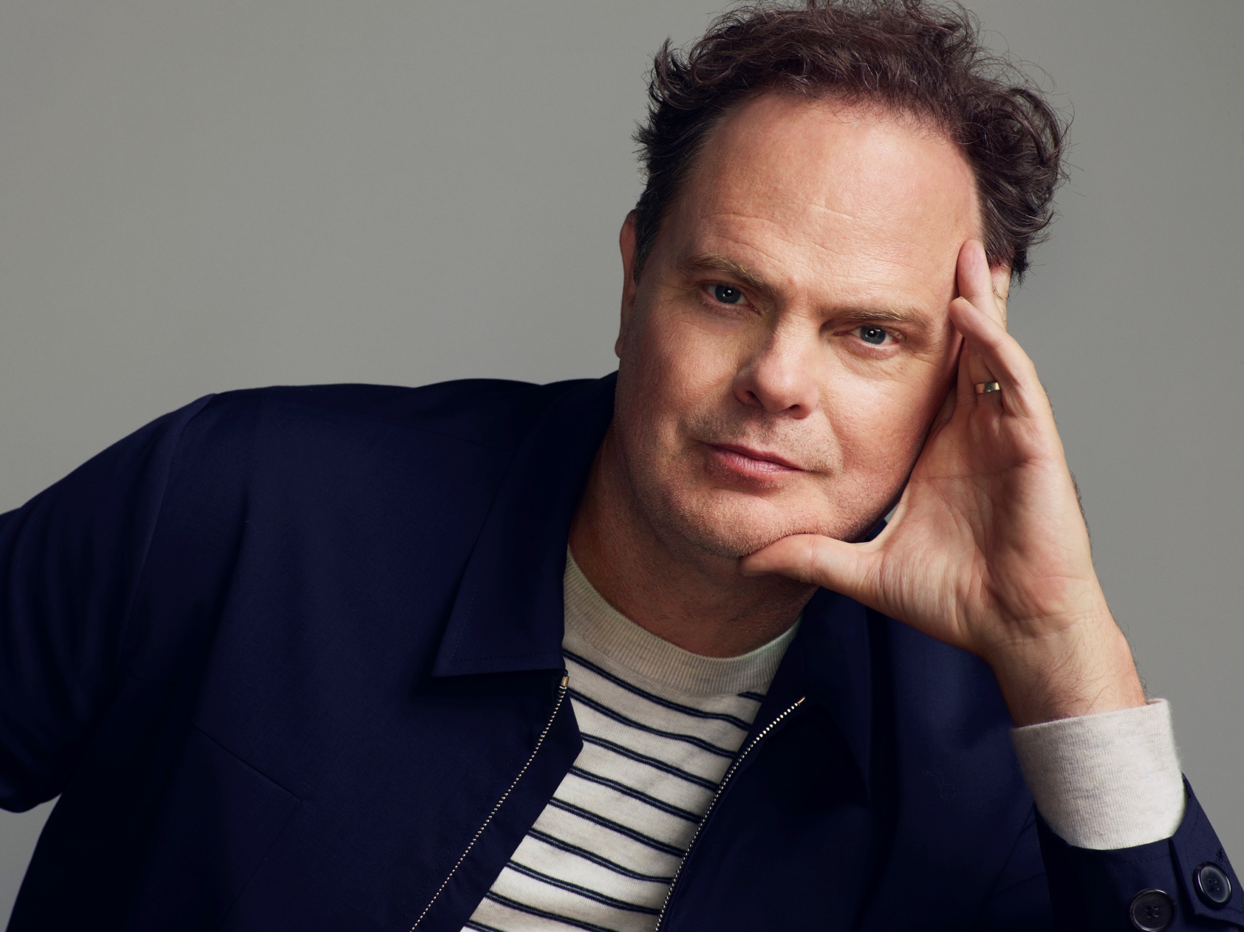 A middle-aged man with fair skin and slightly tousled dark brown hair poses in a relaxed manner. He rests his head on his hand with his fingers lightly touching his temple, looking directly into the camera with a calm and confident expression. He has light stubble on his face and is wearing a navy-blue jacket over a cream and navy striped sweater. The background is a simple, neutral gray, emphasizing his face and expression.