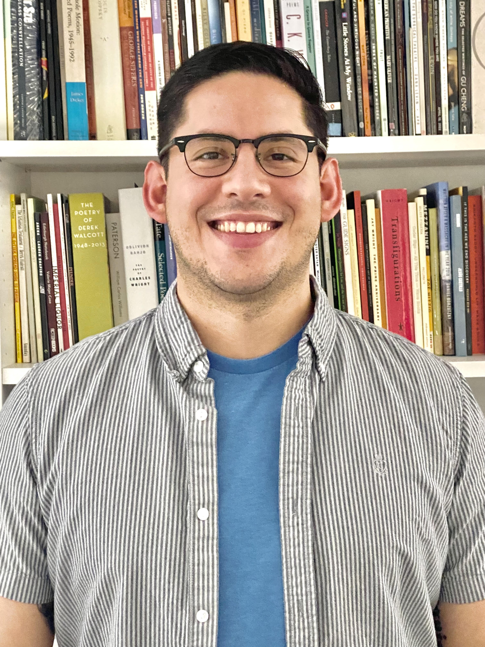 Esteban Rodriguez – Author Photo A person wearing glasses, a blue t-shirt, and a striped button-up shirt stands smiling in front of a bookshelf filled with books.