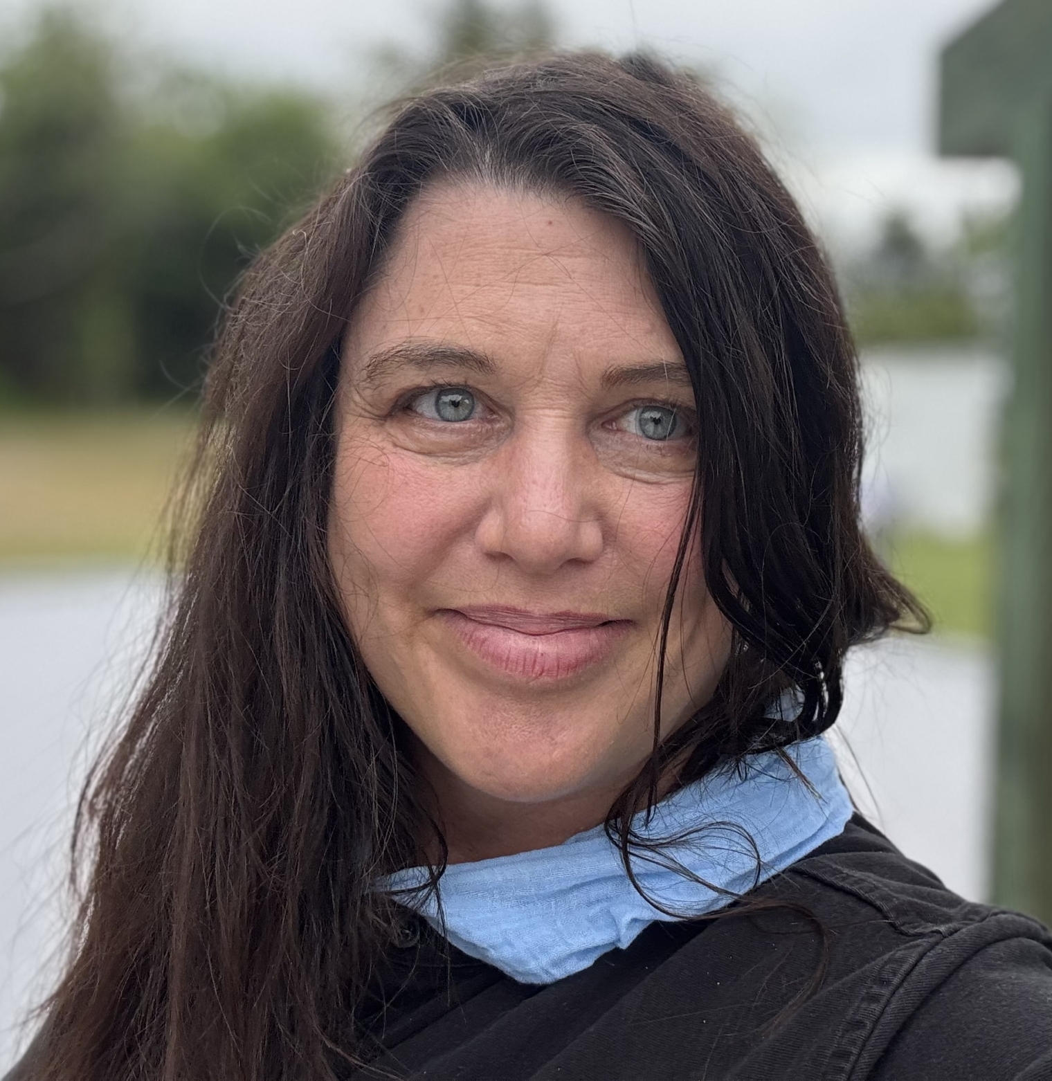 IMG_5688 A woman with long dark hair stands outdoors near a large bell, wearing a black shirt and a blue face mask under her chin. Trees and greenery are visible in the background.