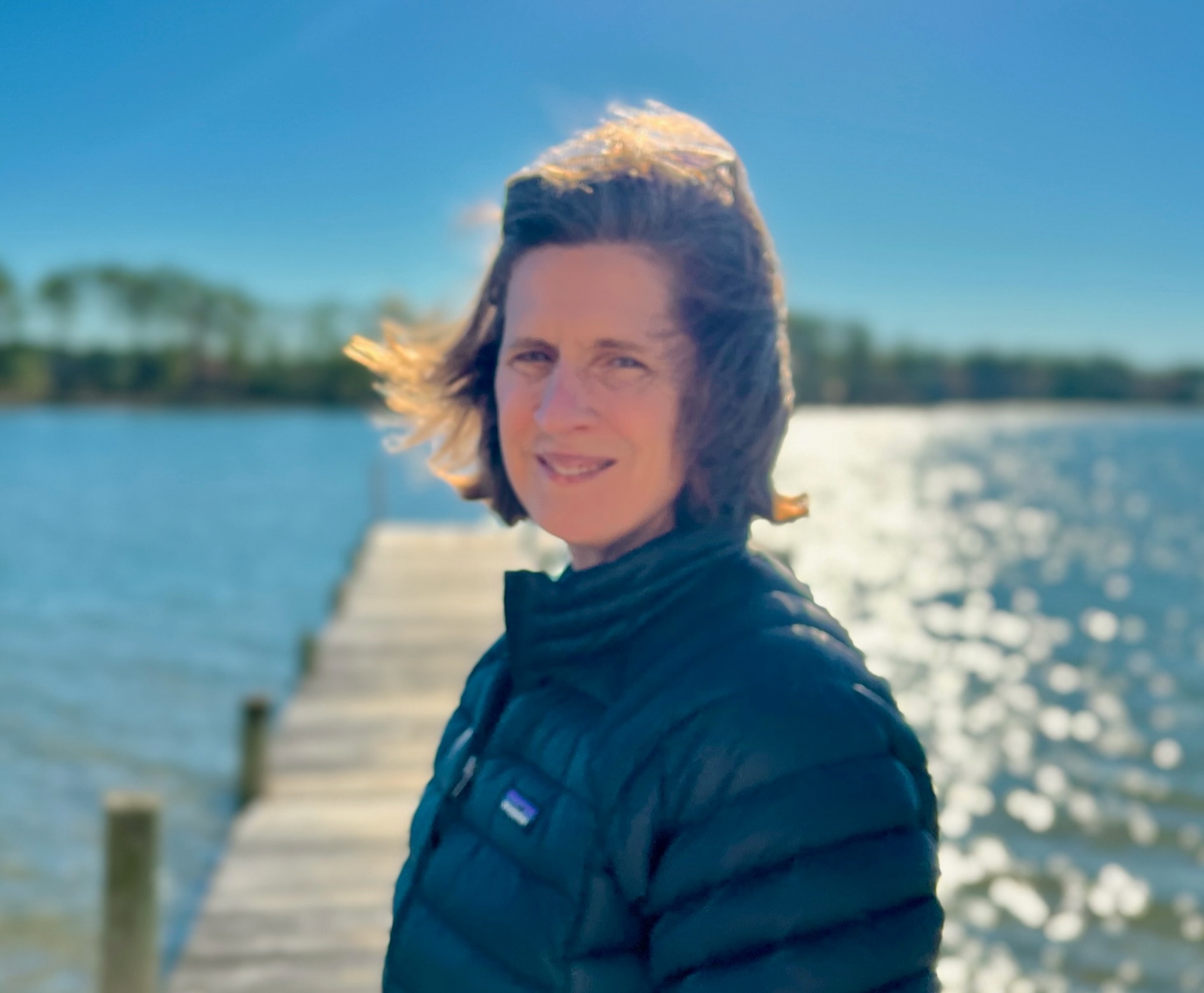 A woman wearing a black jacket stands on a wooden dock by a sunny lake, with trees and a clear blue sky in the background.