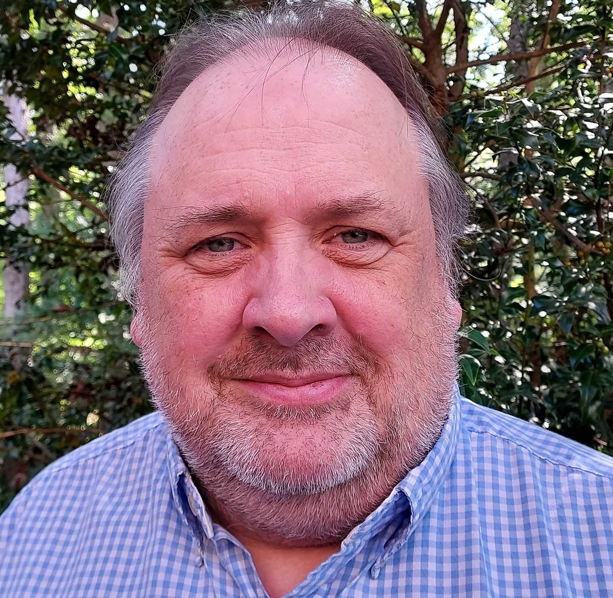 A middle-aged man with light skin, gray hair, and a beard, wearing a blue checkered shirt, smiling outdoors in front of green foliage.
