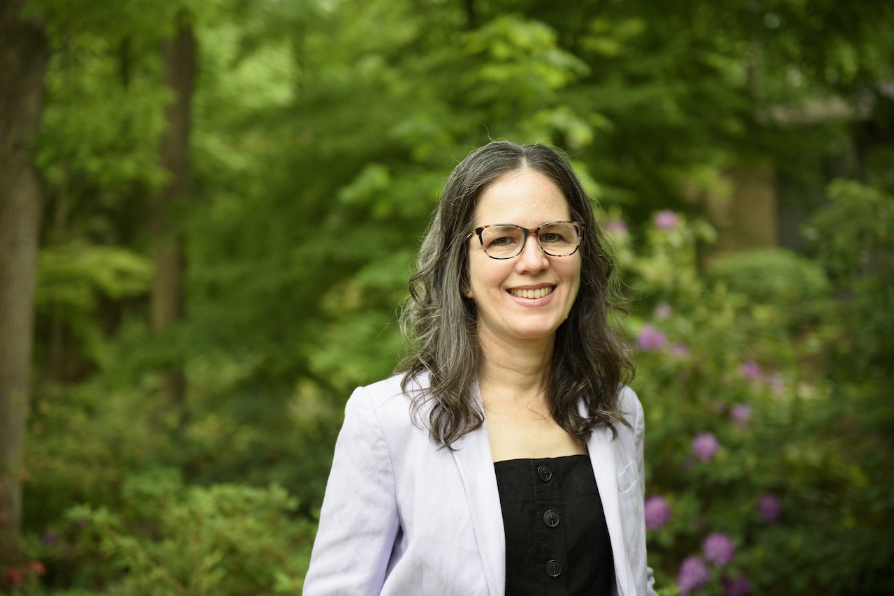 A woman with glasses and long brown hair, wearing a light-colored blazer and black top, stands outdoors in a lush, green garden.