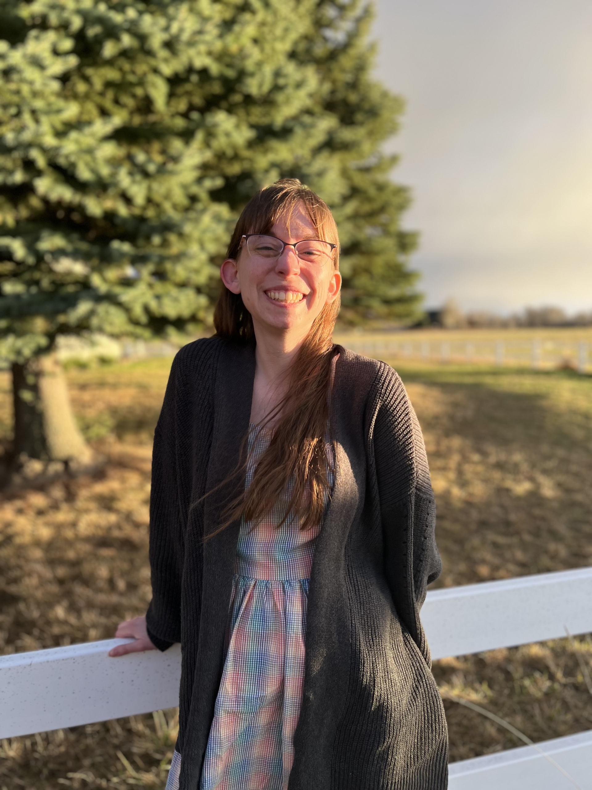 A woman with long brown hair and glasses smiles while leaning against a white fence, with trees and sunlight in the background.