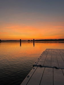 A wooden dock extends into a calm lake under an orange and pink sunset sky, with a few posts visible in the water.