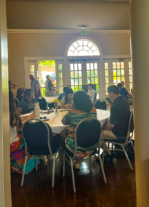 A group of people sit and talk around a round table in a sunlit room with large windows and an arched doorway in the background.