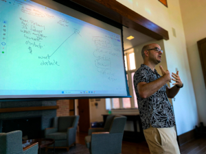 A man stands in front of a projected whiteboard presenting notes and diagrams to an audience in a well-lit room with chairs and large windows.