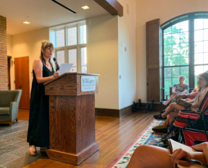 A woman stands at a podium labeled "St. Mary's College" reading from a paper to an audience seated in a well-lit room.
