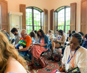 A group of adults sits in a classroom with large windows, focused on writing or taking notes during a workshop or seminar.