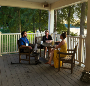 Three people sit and talk at a wooden table on a covered porch overlooking trees and a lake, with drinks on the table.