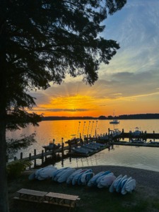 Kayaks and paddleboards are lined up on the shore near a dock at sunset, with boats on the water and a tree framing the vibrant sky.