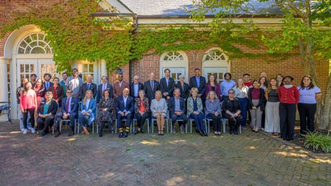 A group photo of approximately 30 people posed outside in front of a brick building with ivy and arched windows. The group is a mix of seated and standing individuals, with trees and a paved area visible in the foreground.