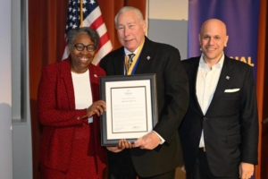 Three people stand together, smiling. The person in the center holds a framed document or award. They are in a room with an American flag and a blue banner in the background.