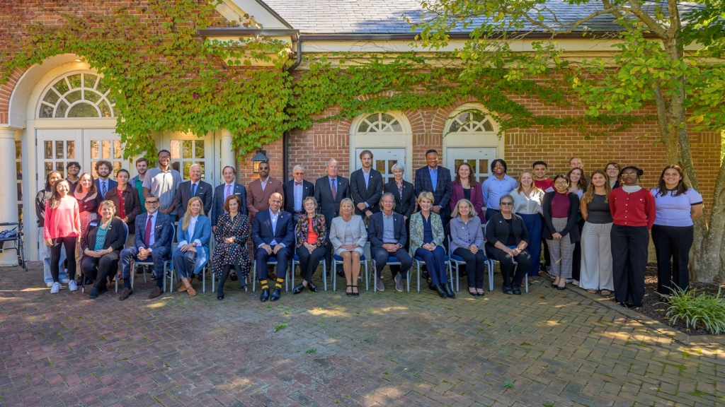A large group of people, dressed in business and casual attire, pose for a group photo outdoors in front of a brick building with greenery.
