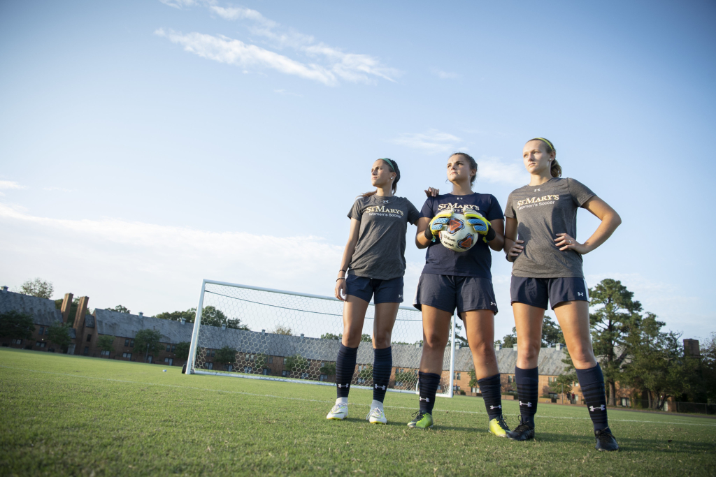 Three female soccer players in uniforms stand on a grassy field in front of a goal, looking into the distance on a clear day.