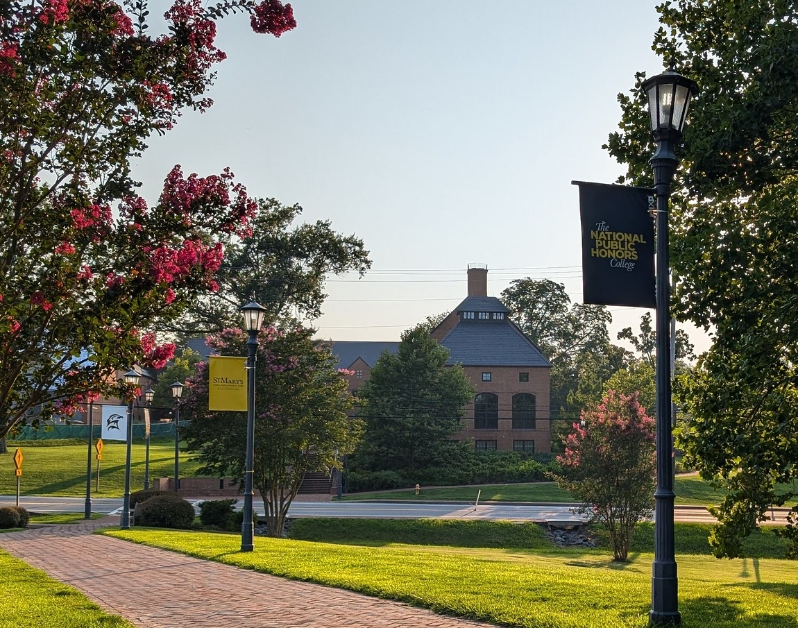A college campus scene with brick buildings, lamp posts with banners, manicured lawns, and blooming trees under clear skies.
