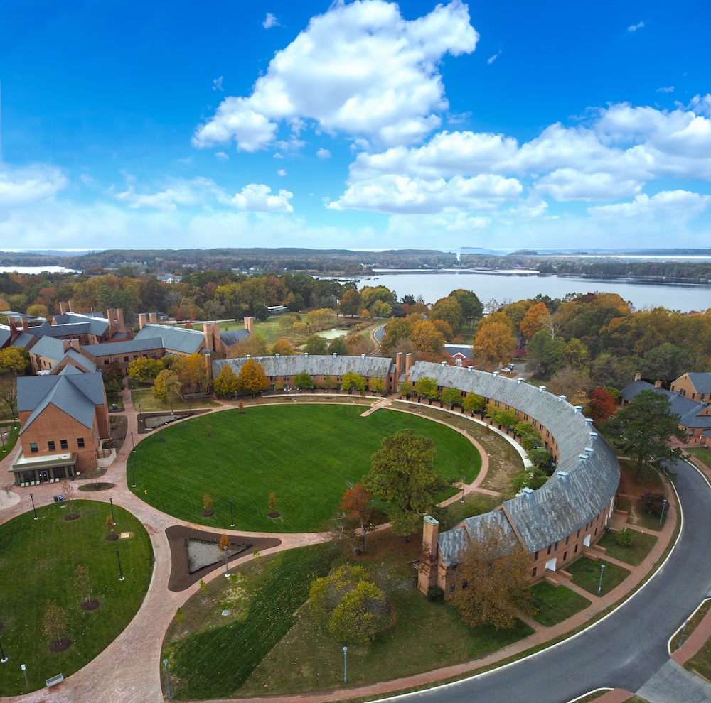 Aerial view of a large, semicircular building surrounding a green lawn, with trees, pathways, other buildings, and a body of water in the background under a blue sky with clouds.