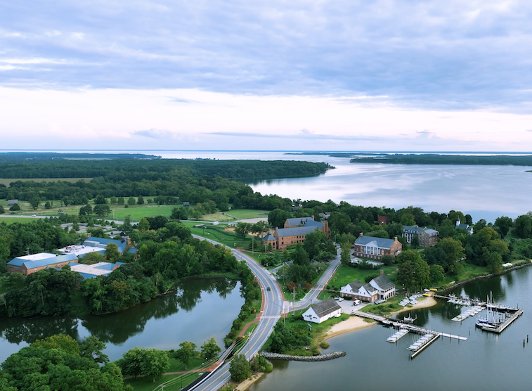 Aerial view of a waterfront village with buildings, a road, marina with boats, trees, and a large body of water under a cloudy sky.
