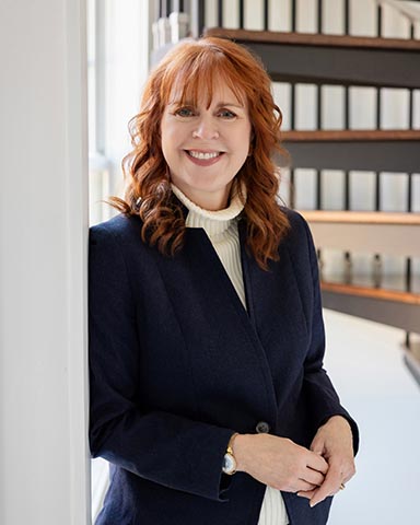 Woman with red hair wearing a navy blazer and white turtleneck stands indoors, smiling, with shelves of binders in the background.