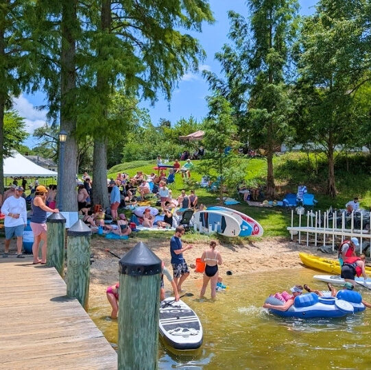 People gather on a grassy hill and sandy beach by a lake, with kayaks, paddleboards, and inflatables in the water on a sunny day.