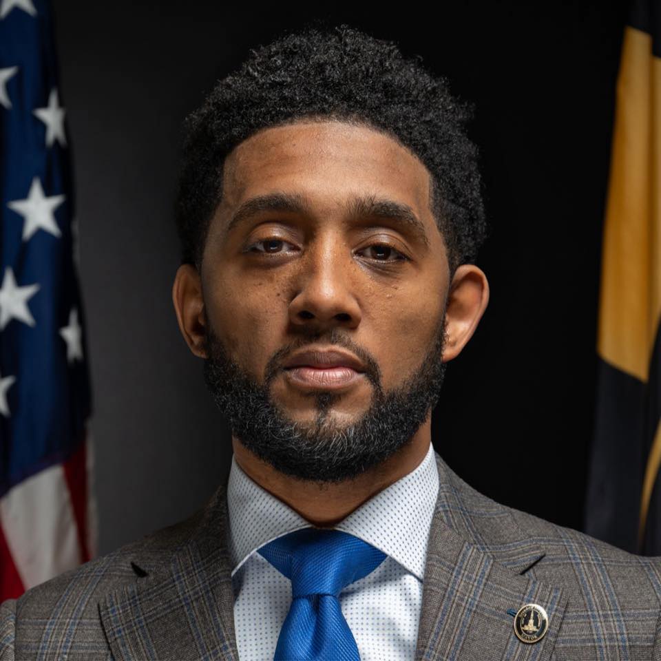 A man in a plaid suit, blue tie, and white shirt stands in front of the U.S. and Maryland flags, looking directly at the camera.