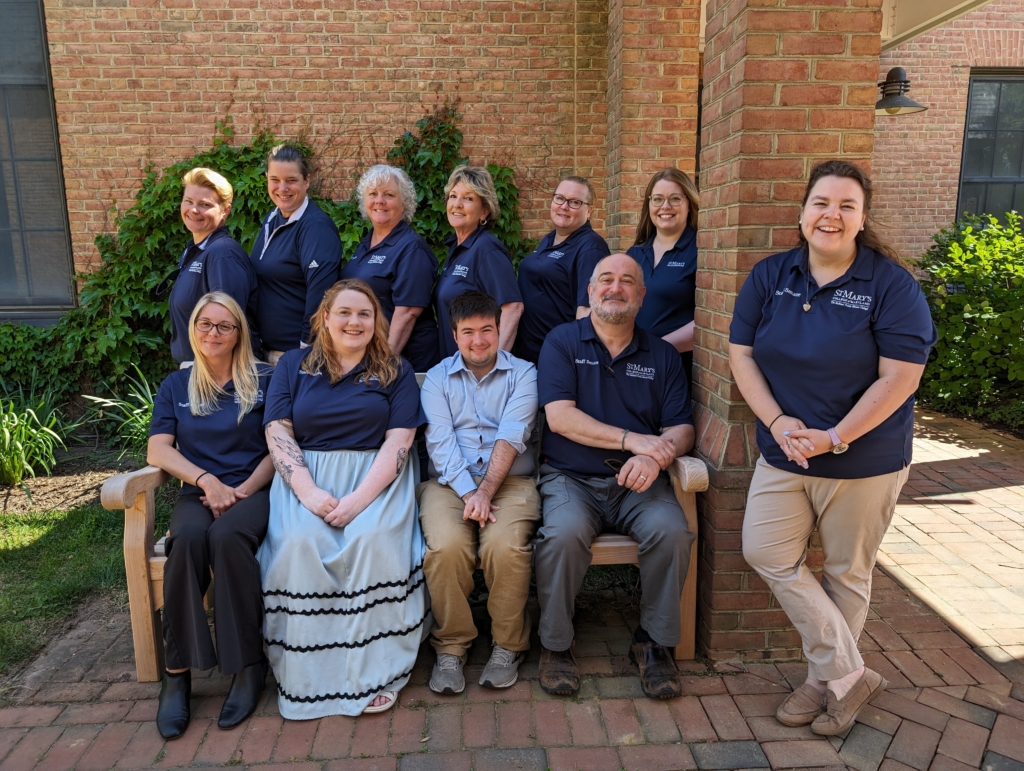 A group of eleven people, wearing matching navy blue shirts, pose together outside a brick building. Some are seated on a bench while others stand behind them, all smiling. Greenery and a brick pathway are visible in the background.