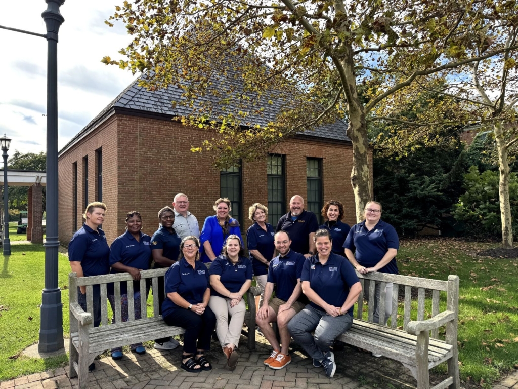 A group of thirteen adults pose together outside on wooden benches in front of a brick building and trees on a sunny day.