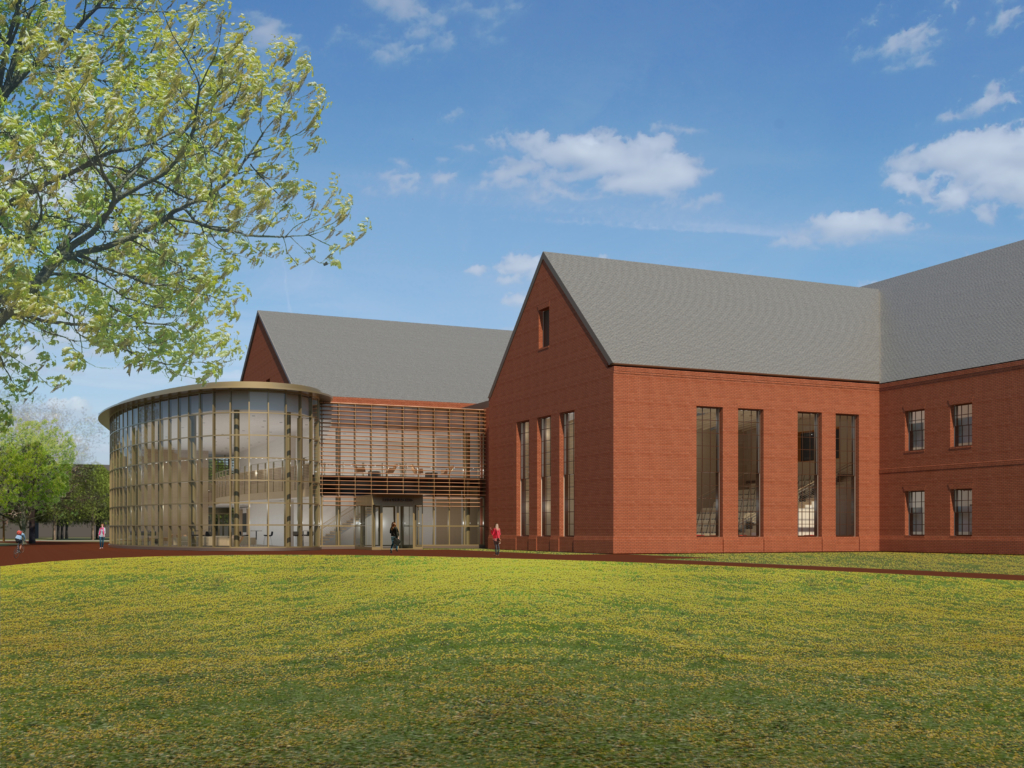 A modern brick building with large windows and a cylindrical glass section, set against a blue sky with scattered clouds. The foreground features a grassy area with a tree on the left. A person walks near the entrance.
