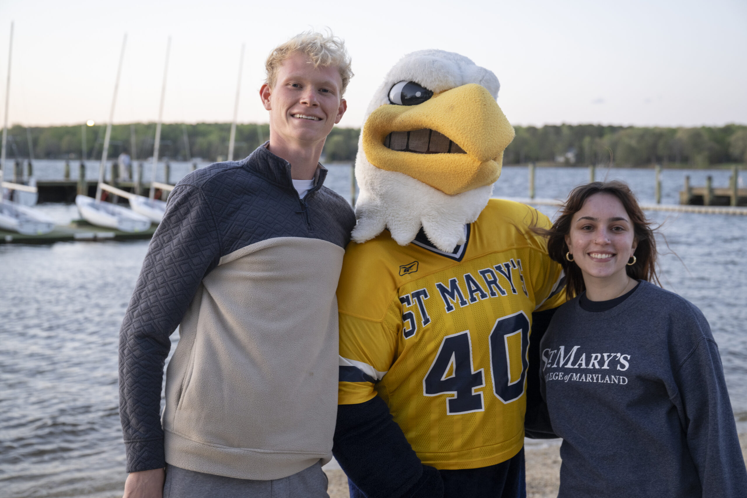 Two students stand on either side of a person in an eagle mascot costume wearing a "St. Mary's 40" jersey, with sailboats and water in the background.