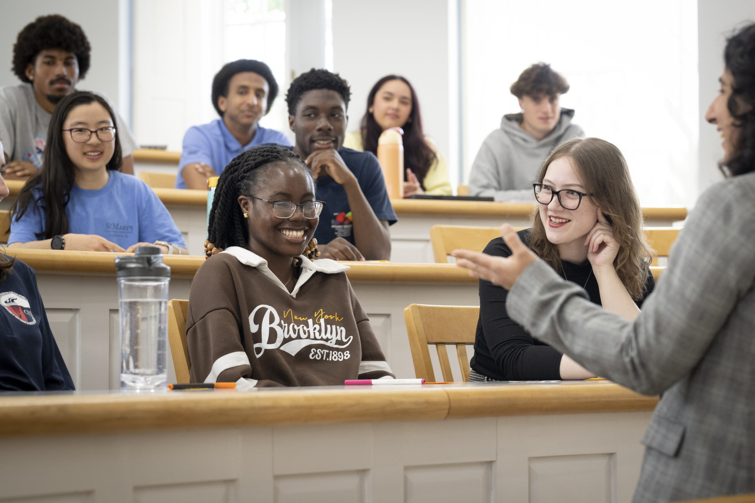 A diverse group of students sits in a lecture hall, attentively listening and smiling as a professor speaks at the front of the classroom.