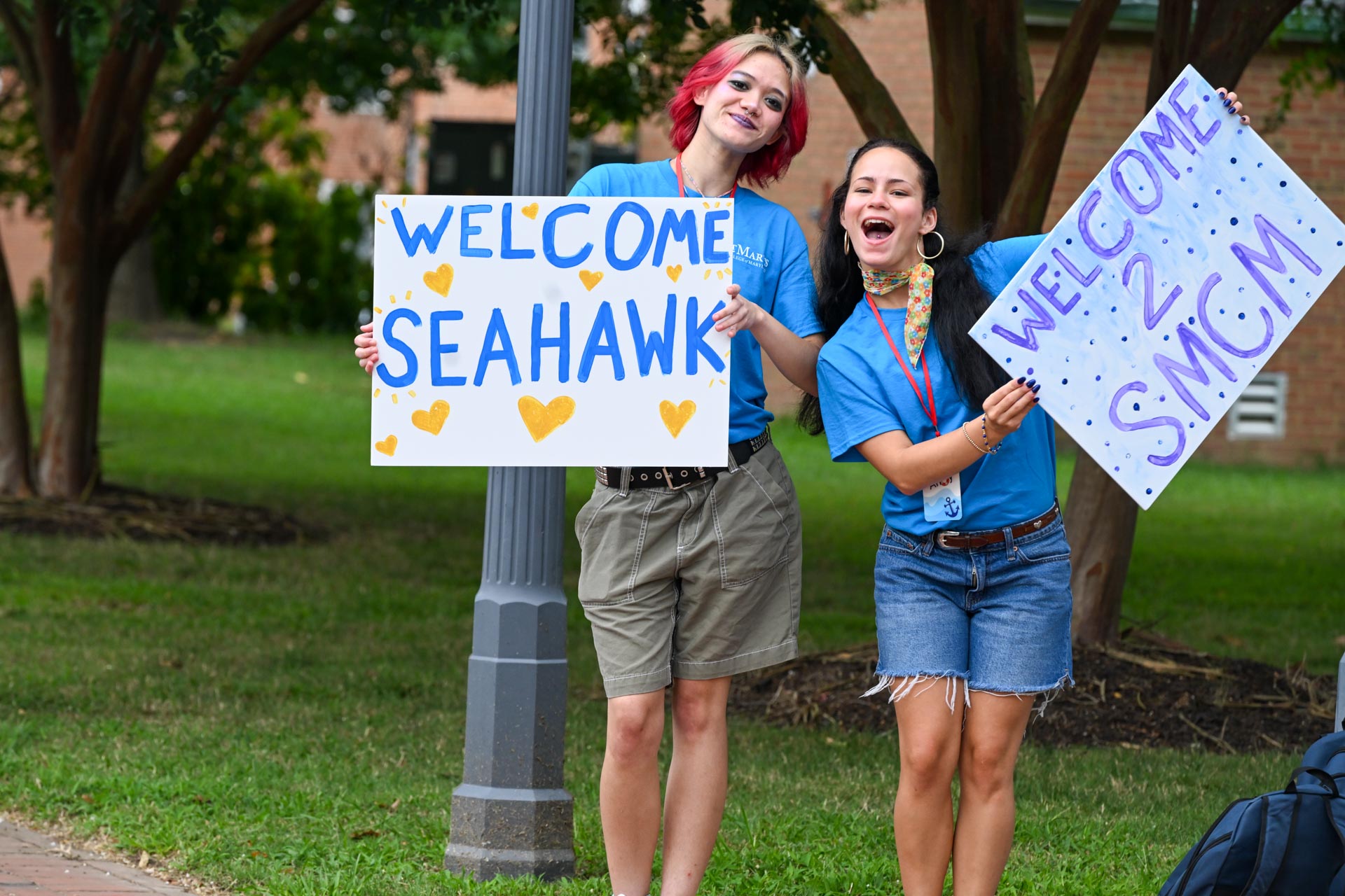 Two people in blue shirts hold welcome signs reading "Welcome Seahawk" and "Welcome 2 SMCM" while standing outside on a grassy campus area.