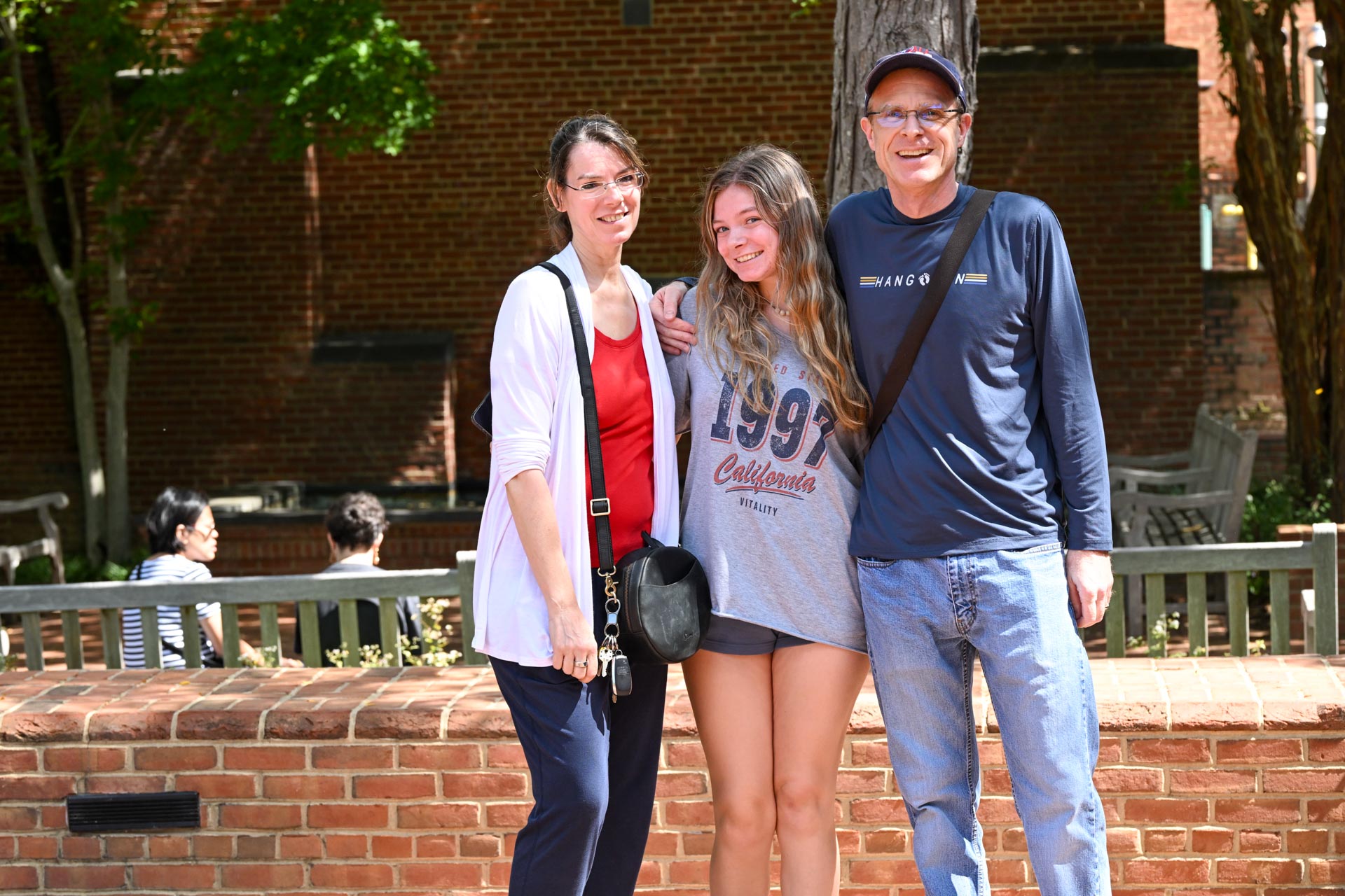 Three people stand together outdoors in front of a brick wall, smiling at the camera. Two adults flank a young woman wearing a gray t-shirt and shorts. Trees and benches are in the background.