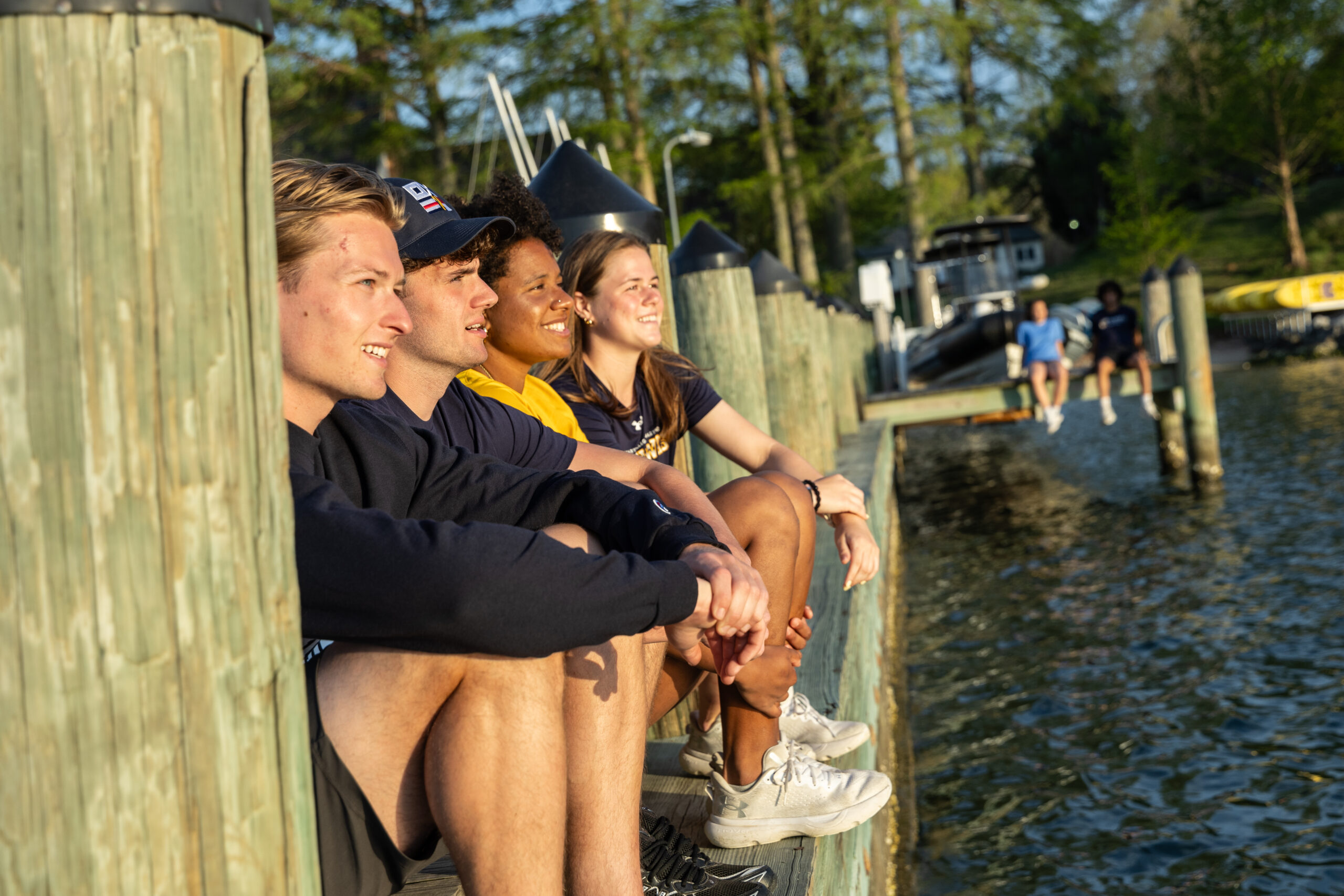 Four young adults sit on a wooden dock by the water, looking out and smiling. Trees and boats are visible in the background.