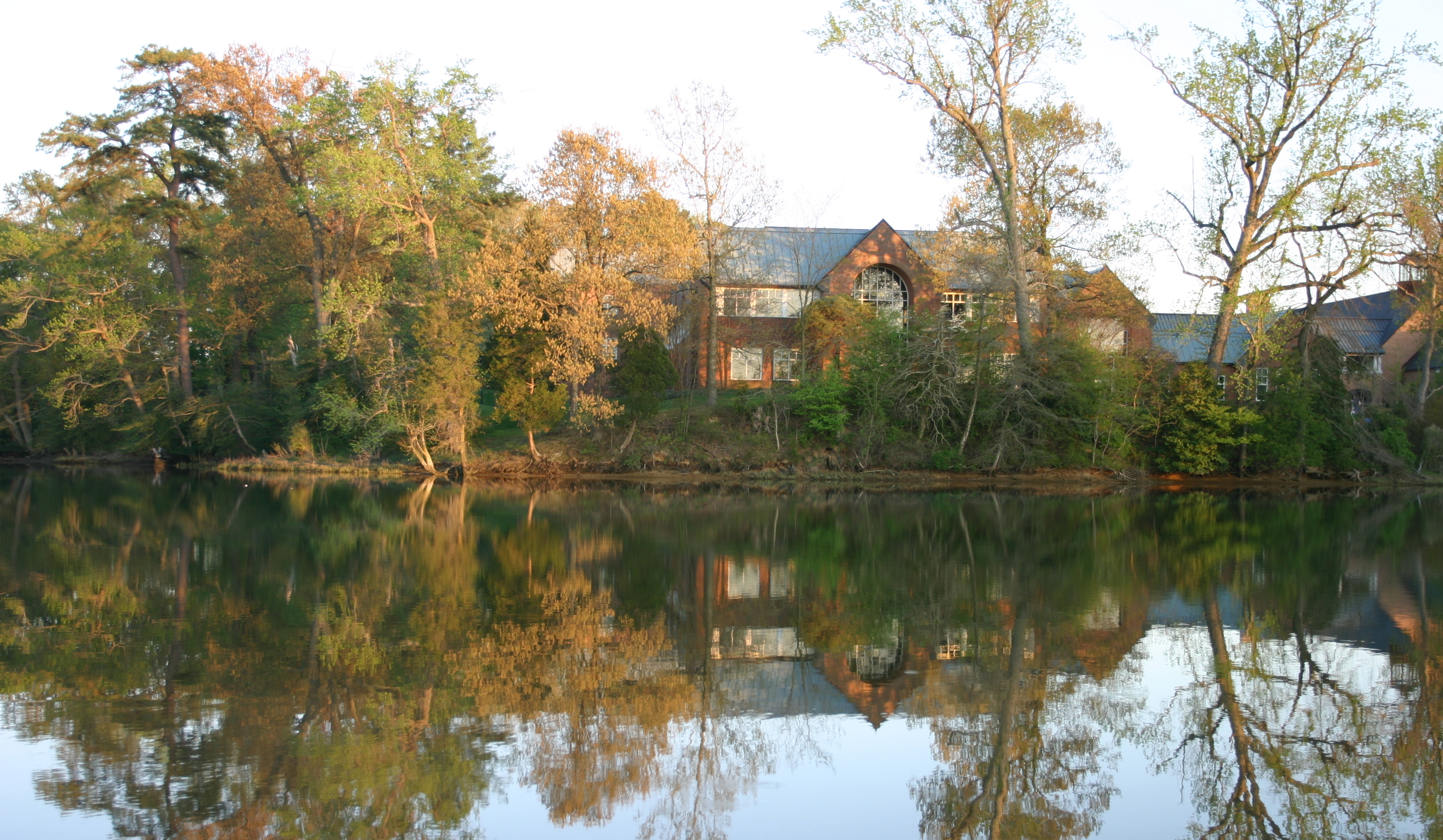 A serene lake reflects a large house surrounded by tall trees with autumn foliage. The house is partially obscured by the trees, and the calm water mirrors the scene, enhancing the tranquil atmosphere.