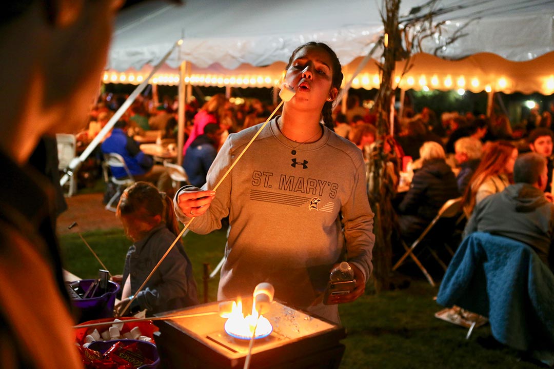 A woman in a St. Mary's sweatshirt roasts a marshmallow over a small flame at an outdoor event with people seated under a large tent in the background.