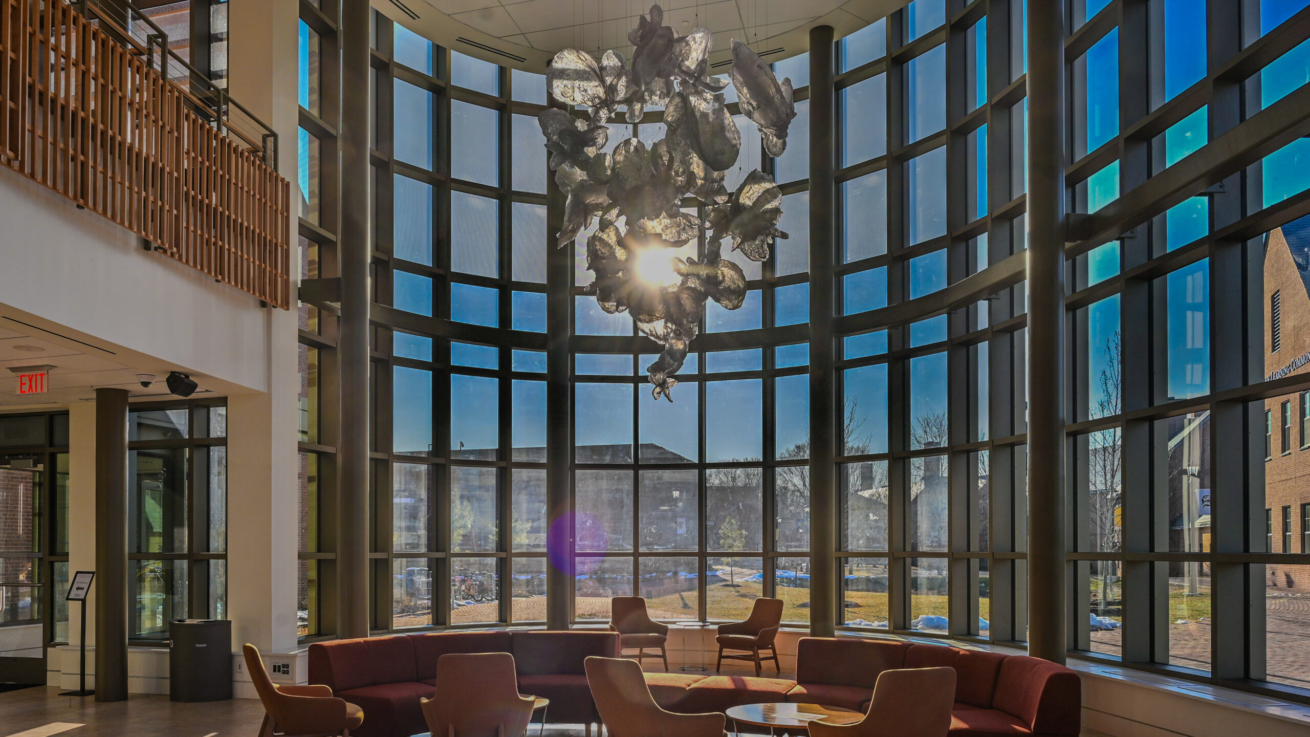 A sunlit facility lobby with curved floor-to-ceiling windows, modern seating, and a large abstract chandelier hanging from the ceiling.