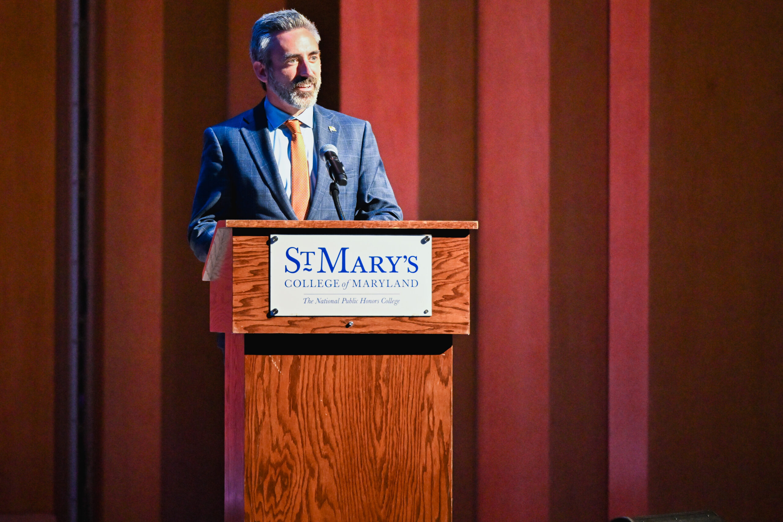 A man in a suit stands at a podium with a microphone, delivering a speech at St. Mary's College of Maryland during an event.