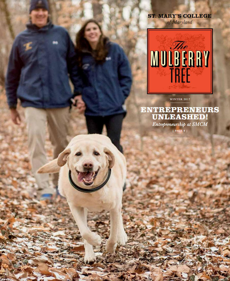 A happy dog runs through a wooded area covered with fallen leaves. In the background, a man and woman in blue jackets hold hands and walk together. The cover text reads "The Mulberry Tree, Winter 2017" and mentions entrepreneurship at St. Mary's College of Maryland.