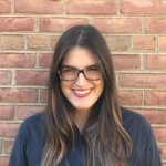 A person with long brown hair and glasses smiles while standing against a brick wall. They are wearing a dark top.
